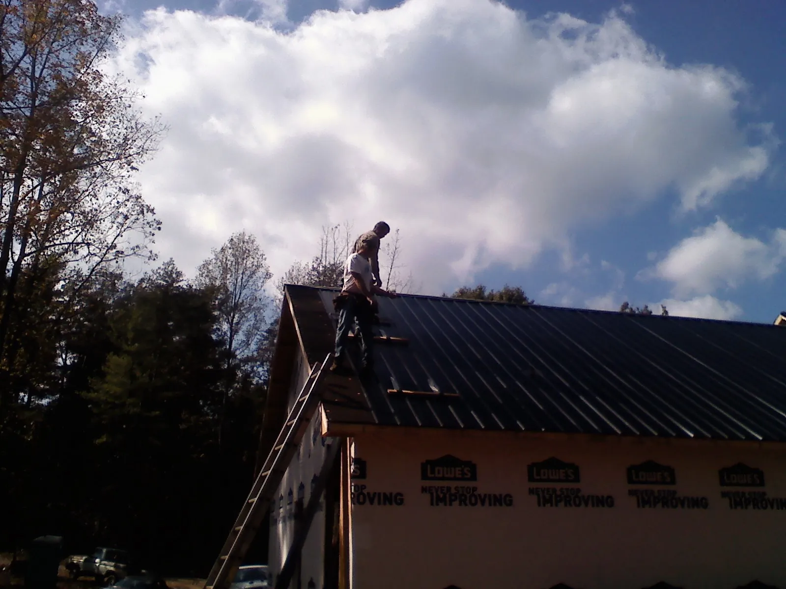 Roofer fastening dark metal panels high against a cloudy sky, from a Roof Inspection job in Massapequa